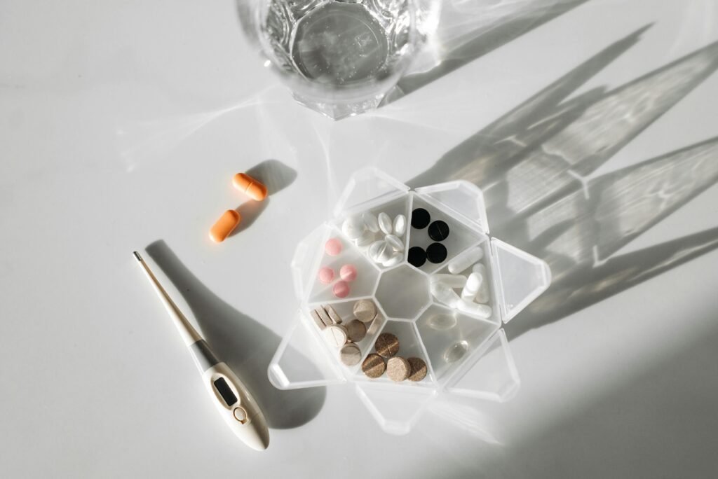 A detailed image showcasing a pill organizer, digital thermometer, and pills with a glass of water on a white surface.
